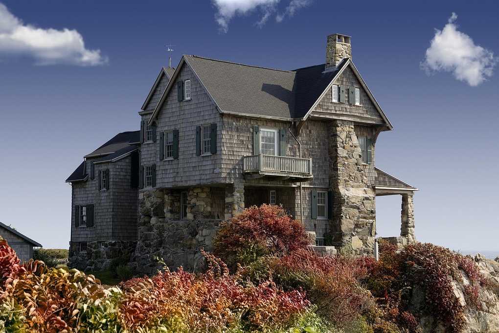Maison sur falaise entourée de buissons colorés, toit en ardoise, façade en pierre et bois, ciel bleu