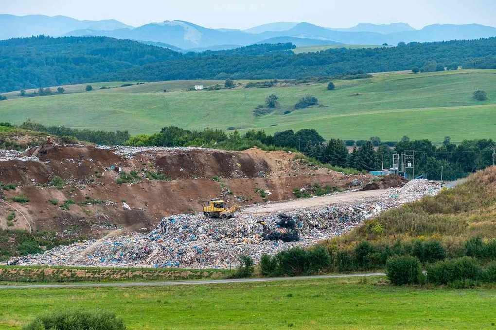 Site de décharge sauvage avec déchets entassés et pelleteuse jaune, paysage agricole en arrière-plan Site de décharge sauvage avec déchets entassés et pelleteuse jaune, paysage agricole en arrière-plan